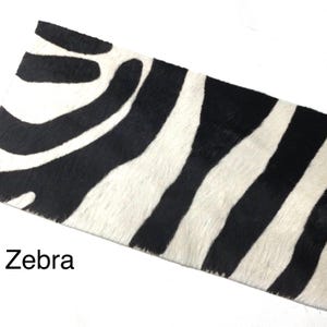 May include: A rectangular piece of animal hide featuring a high-contrast black and white zebra stripe pattern. The surface has a visible hair texture throughout the material. The word Zebra is printed in black text in the lower left corner.