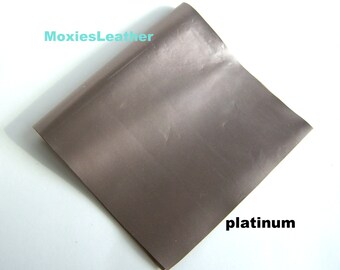 May include: A sheet of platinum-colored leather, folded over, with the word "platinum" printed in black. The text "MoxiesLeather" is printed in light blue in the upper left corner. The leather has a metallic sheen.