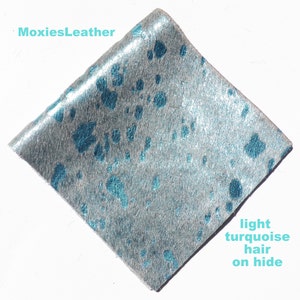 May include: A close-up of a piece of light turquoise cowhide leather with a silver metallic finish. The leather has a soft, fuzzy texture and is perfect for crafting.