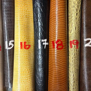 Può includere: Sette rotoli verticali di materiale testurizzato con motivi in rilievo. I colori da sinistra a destra sono beige, marrone scuro, giallo, nero, marrone chiaro, oro metallizzato e marrone scuro. Ogni rotolo è etichettato con i numeri da 14 a 20.