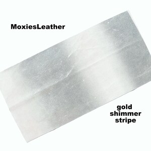 Puede incluir: Una tela blanca y plateada con una franja brillante dorada. La tela se llama "MoxiesLeather".