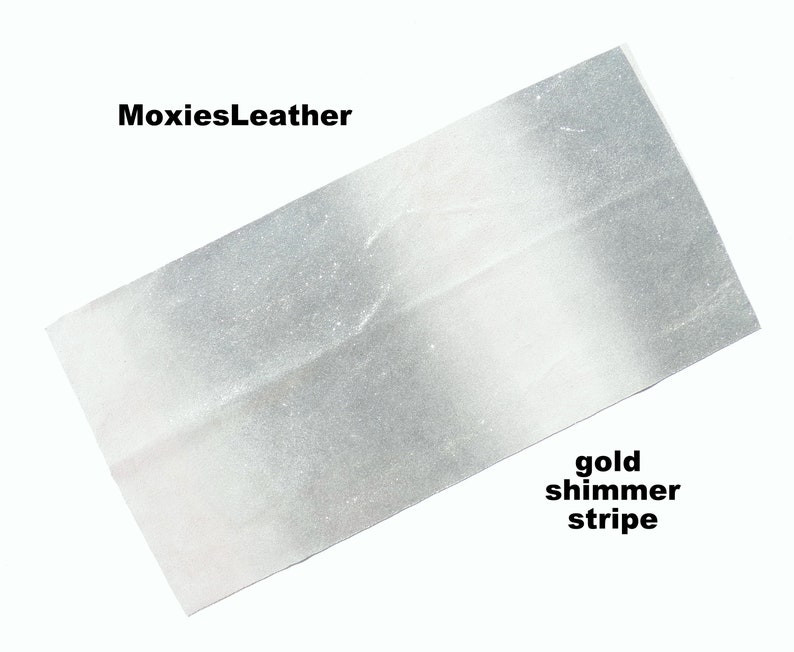 Puede incluir: Una tela blanca y plateada con una franja brillante dorada. La tela se llama "MoxiesLeather".