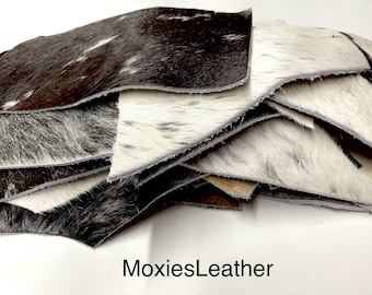 May include: A stack of cowhide leather pieces in various shapes and sizes. The leather has a natural, textured appearance with a mix of black, white, and brown colors. The text "MoxiesLeather" is visible at the bottom.