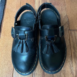 May include: A pair of black leather Dr. Martens sandals with a buckle and tassel detail. The sandals have a black leather strap that goes around the ankle and a black leather buckle.