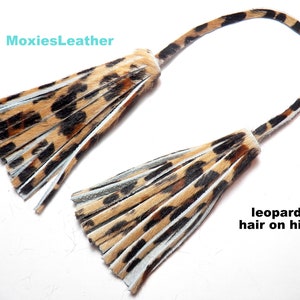 Op de afbeelding: Twee bruine en zwarte luipaardprint kwastjes met witte bies, bevestigd aan een bruin en zwart luipaardprint lederen koord. "MoxiesLeather" en "luipaardhaar op leer" staan in de afbeelding.