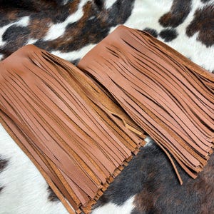 May include: Two brown leather fringe pieces, suitable for crafting or decoration. The fringe is finely cut and hangs in neat rows. The leather has a warm, natural tone, and the pieces are displayed against a cowhide pattern.