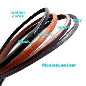 Könnte beinhalten: Fünf Lederbänder in verschiedenen Farben: Cognac, Braun, Grau, Schwarz und Schwarz. Die Bänder sind in einem Halbkreis angeordnet, wobei das Cognac-Band oben liegt. Der Text "MoxiesLeather" ist in der unteren rechten Ecke des Bildes sichtbar.