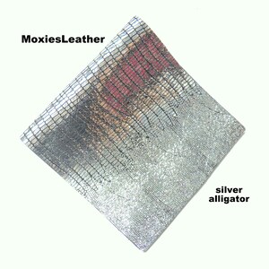 Puede incluir: Un cuero de piel de cocodrilo plateado con un patrón texturizado. MoxiesLeather está impreso en la esquina superior izquierda.