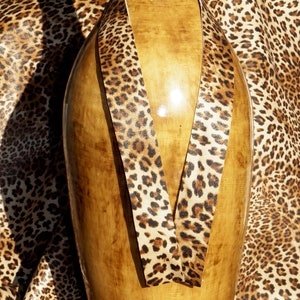 Op de afbeelding: Een bruin leren band met een luipaardprint. De band is om een ​​houten vaas gewikkeld. De tekst "MoxiesLeather" is zichtbaar onderaan de afbeelding.