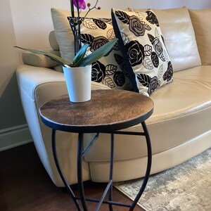 May include: A small, round, wooden table with a black metal base. The table is next to a white couch with floral patterned pillows. A potted orchid with pink flowers is on the table.