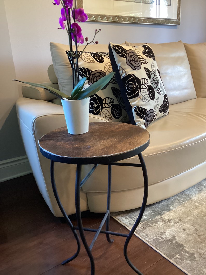 May include: A small, round, wooden table with a black metal base. The table is next to a white couch with floral patterned pillows. A potted orchid with pink flowers is on the table.