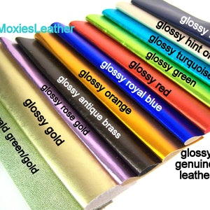 May include: A close-up of ten different colours of leather swatches. The swatches are arranged in a fan shape, with the darkest colour at the top and the lightest colour at the bottom. The colours are: glossy navy, glossy hint of gold, glossy turquoise, glossy green, glossy royal blue, glossy red, glossy orange, glossy antique brass, glossy rose gold, and glossy gold. The bottom swatch is labelled "emerald green/gold".