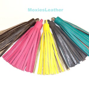 Puede incluir: Cinco borlas de cuero en diferentes colores: marrón, rosa, amarillo, gris y verde azulado. Las borlas están dispuestas en forma de abanico, con la borla marrón en la parte superior y la borla verde azulada en la parte inferior.