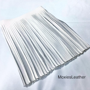 May include: A white leather fringe piece, likely for crafting or fashion. The fringe is made of many thin, parallel strips of white leather. The texture is smooth and the overall shape is rectangular. The brand name "MoxiesLeather" is visible in the bottom right corner.