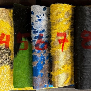 May include: Nine rectangular swatches of textured hair-on-hide leather arranged side-by-side, each marked with a red number from one to nine. The swatches feature various colors and patterns, including solid yellow, black, green, and spotted designs.
