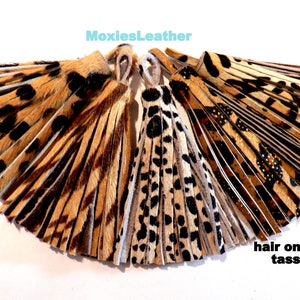 May include: A set of five brown, black, and white hair on hide tassels with a leopard print pattern. The tassels are arranged in a fan shape. The text "MoxiesLeather" and "hair on hide tassels" is visible in the image.