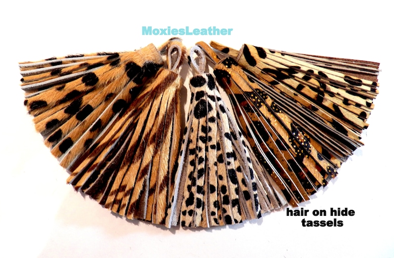 May include: A set of five brown, black, and white hair on hide tassels with a leopard print pattern. The tassels are arranged in a fan shape. The text "MoxiesLeather" and "hair on hide tassels" is visible in the image.