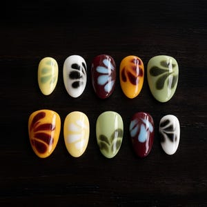 May include: A set of ten almond-shaped press-on nails featuring various glossy base colors including yellow, white, burgundy, orange, and sage green, each decorated with a painted floral-like pattern in contrasting shades of black, white, or blue.