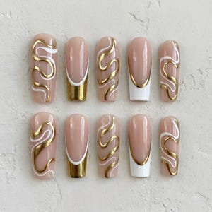 May include: Set of ten artificial nails in a neutral beige hue, adorned with gold and white wavy designs. Some nails feature a gold or white French tip. The nails are designed for a stylish manicure.
