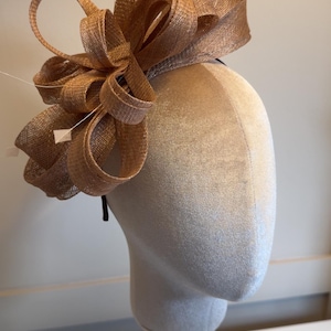 May include: A metallic bronze fascinator headpiece featuring a large, multi-looped bow made of stiffened, woven fabric. The headpiece is adorned with thin, delicate white feather accents and is mounted on a thin, dark-coloured headband.