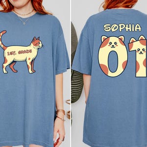 Puede incluir: Una camiseta azul con un gráfico de un gato y el texto 1ST GRADE en la parte delantera. La parte trasera muestra el nombre SOPHIA sobre el número 01, estilizado con orejas de gato y un patrón de manchas, a juego con el gráfico del gato.
