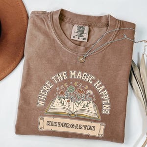 Op de afbeelding: Een bruin t-shirt met korte mouwen en een afbeelding van een open boek waar bloemen uit groeien, omringd door sterren en manen. De tekst "WHERE THE MAGIC HAPPENS" staat in een boog erboven en "KINDERGARTEN" op een banner onder het boek.
