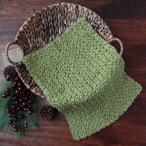 May include: A green crocheted square blanket rests in a woven basket with handles. The blanket is a solid color and has a textured appearance. Decorative pinecones, faux berries, and greenery are arranged around the basket on a wooden surface.