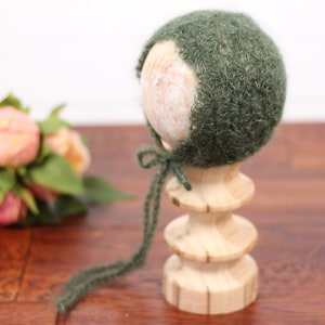 May include: A dark green knitted baby bonnet with a tie closure, displayed on a wooden stand. The bonnet is made of a soft, textured yarn. Pink and peach flowers are in the background.