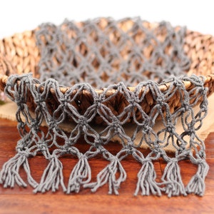 May include: A woven wicker basket with handles, holding a gray macrame scarf with fringe. The basket sits on a tan fabric, against a dark wood surface. Pink flowers are visible in the background.