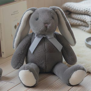 May include: A plush stuffed rabbit toy with dark grey fur, long ears with cream-coloured inner linings, and a light grey ribbon tied around its neck. The rabbit has cream-coloured patches on its feet and a small, dark nose, sitting in a seated position.