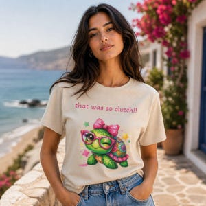 Può includere: T-shirt beige con la scritta "that was so clutch!!" sopra una tartaruga dei cartoni animati con occhiali e fiocco rosa. La tartaruga è verde, rosa e gialla con stelle intorno. La maglietta è girocollo.