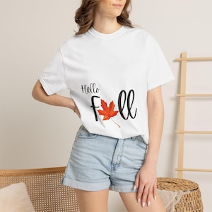 Può includere: Una t-shirt bianca a maniche corte con la scritta nera Hello Fall, in cui la lettera a è sostituita da una grafica di una foglia d'acero arancione e rossa. La maglietta è abbinata a pantaloncini di jeans azzurri con orlo risvoltato.