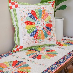 May include: A white quilted table runner with a colorful patchwork design featuring a repeating pattern of pinwheels. The runner has a green and white border and is accented with a matching pillow.