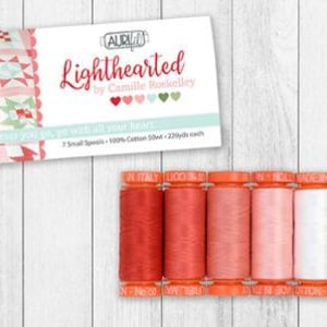 May include: A set of seven spools of thread in shades of red, pink, white, green, and brown. The spools are labeled "Aurifil" and "Lighthearted by Camille Roskelley". The text "Wherever you go, go with all your heart" is also visible on the packaging.