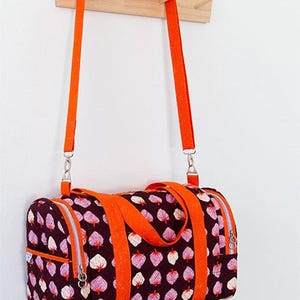 May include: A cylindrical duffle bag with a repeating pattern of pink and white leaf-like shapes on a dark burgundy background. The bag has bright orange straps and a silver zipper. The text "Lucy Duffle" is at the top.