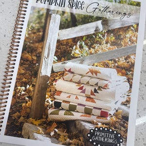 May include: A spiral-bound instructional booklet titled Pumpkin Spice Gatherings featuring a stack of folded quilts with autumn-themed patterns. The cover displays a photograph of the quilts resting against a wooden fence amidst fallen yellow leaves.