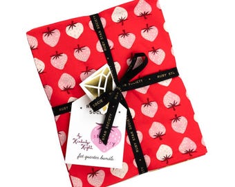 May include: A red fat quarter bundle featuring a strawberry pattern. The fabric is tied with a black ribbon printed with "Ruby Star Society." A tag with a strawberry graphic and text "by Kimberly Kight" is attached.