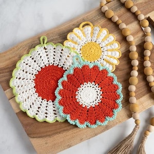 May include: Three colorful, crocheted flower-shaped pot holders. One is white with a red center and green trim, another is red with a white center and blue trim, and the third is yellow with white petals. A wooden bead garland is in the background.