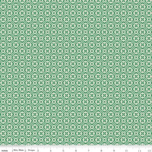 May include: A green fabric featuring a repeating geometric pattern of white diamonds and four-petaled floral shapes. A white ruler at the bottom edge shows a scale of 12 inches, with the text Riley Blake Designs printed on the left side.
