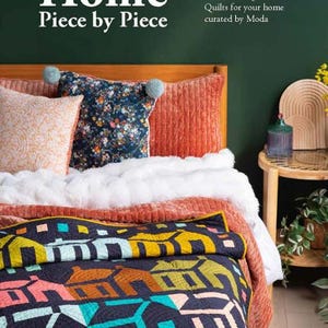 May include: A book titled Home Piece by Piece: Twelve Fat Quarter Quilts for your home curated by Moda. The cover features a bed with a dark blue quilt displaying a colorful house pattern, a white throw, orange pillows, and a floral pillow.