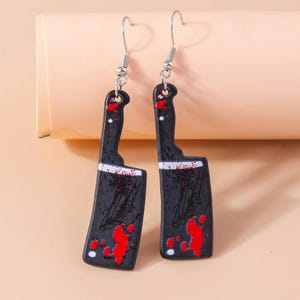 May include: A pair of novelty earrings featuring a cleaver design. The earrings are black with white accents and red splatters, hanging from silver-colored fish hook ear wires. The earrings are a unique accessory.
