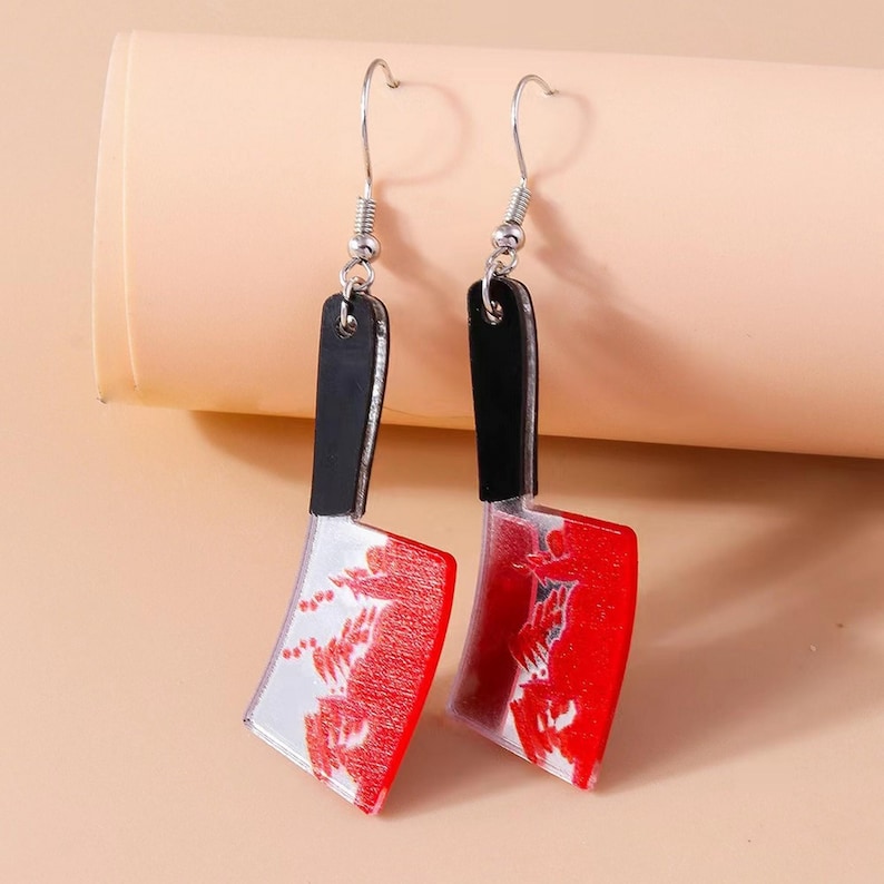May include: A pair of novelty earrings shaped like cleavers. The earrings feature a black handle, a silver hook, and a blade with a red and white design. The earrings are designed to look like a bloody butcher knife.