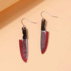 May include: A pair of novelty earrings shaped like miniature knives. The blades are red with the text "KILLING IT" in white, and the handles are black. The earrings hang from silver-colored fish hook ear wires.
