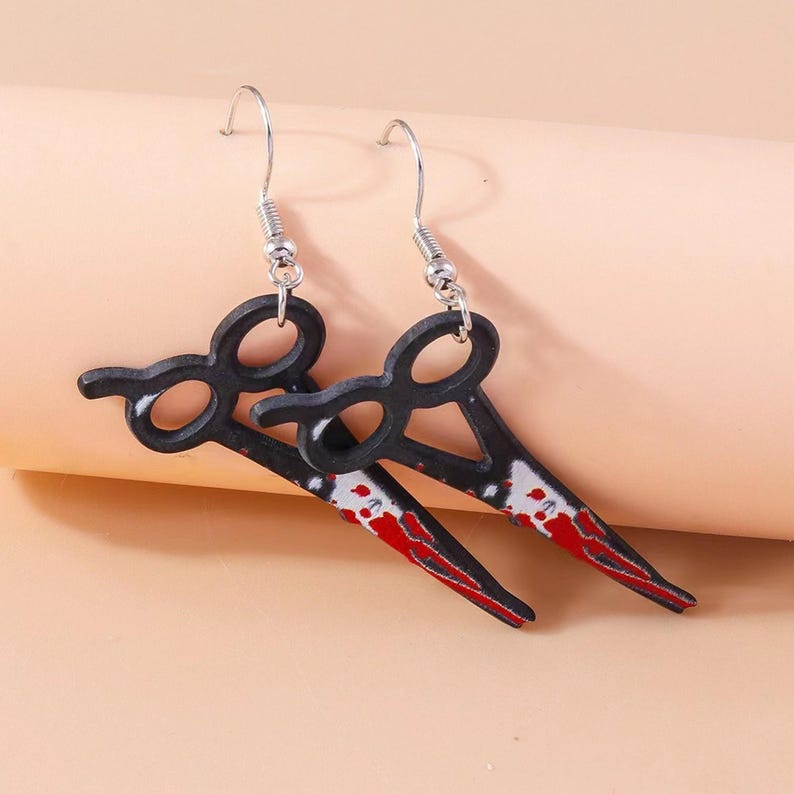 May include: A pair of novelty earrings featuring a scissor design. The black scissors have a red and white blood splatter design on the blades. The earrings hang from silver-colored fish hook ear wires.