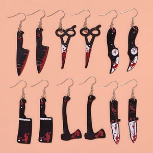 May include: A collection of novelty earrings featuring various weapon designs. The earrings include black and red butcher knives, cleavers, axes, and scissors, all with a blood-splatter effect. The earrings are suspended from silver-colored fish hook ear wires.