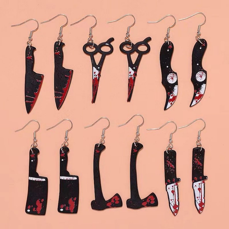 May include: A collection of novelty earrings featuring various weapon designs. The earrings include black and red butcher knives, cleavers, axes, and scissors, all with a blood-splatter effect. The earrings are suspended from silver-colored fish hook ear wires.