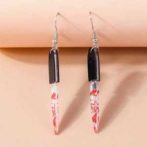 May include: A pair of novelty earrings shaped like miniature knives. Each earring features a black handle and a clear blade with red splatter details, suspended from silver-colored fish hook ear wires. The earrings are designed to resemble a bloody knife.