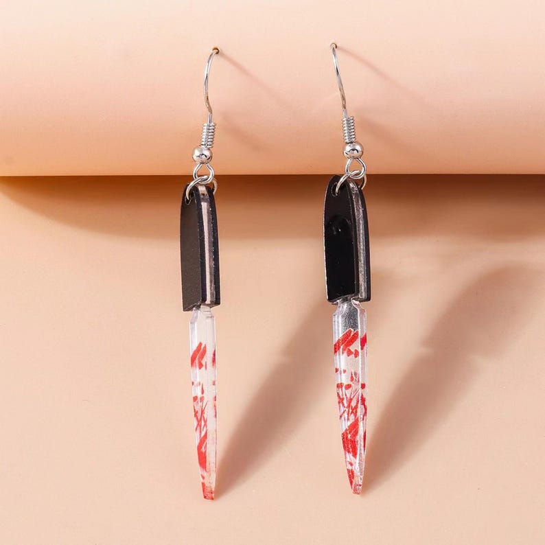 May include: A pair of novelty earrings shaped like miniature knives. Each earring features a black handle and a clear blade with red splatter details, suspended from silver-colored fish hook ear wires. The earrings are designed to resemble a bloody knife.