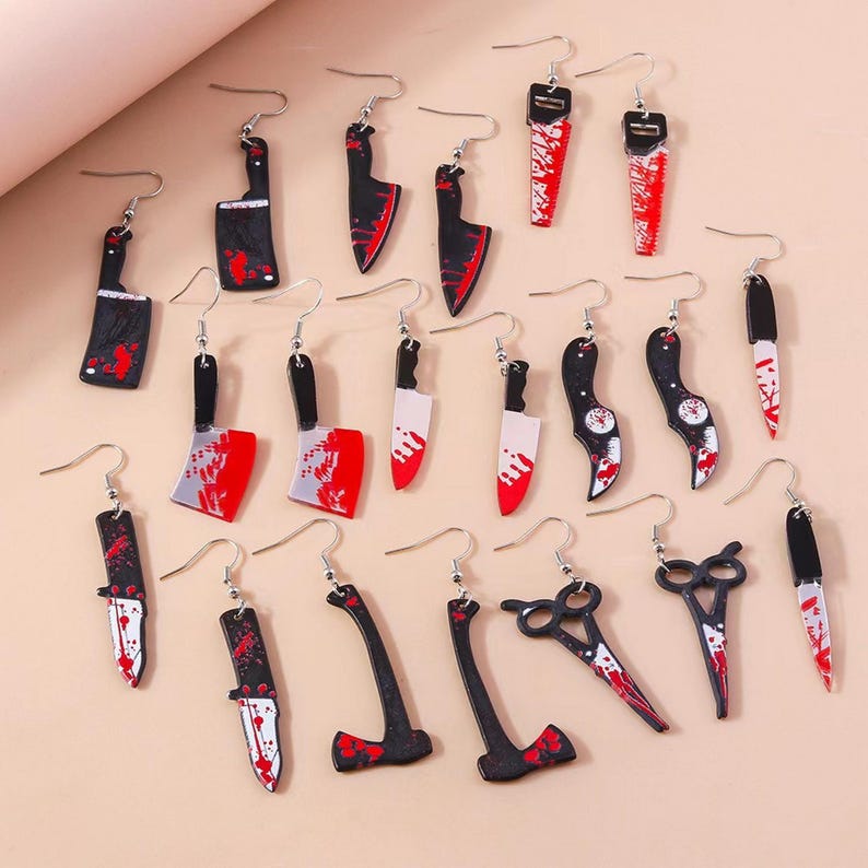 May include: A collection of novelty earrings in various shapes, including knives, cleavers, saws, and scissors. The earrings are black and silver with red accents, designed to resemble blood. Each earring has a silver hook.