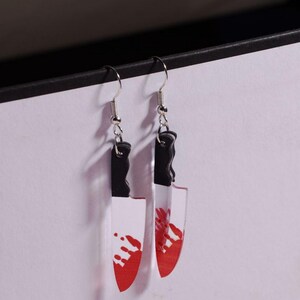 May include: A pair of novelty earrings shaped like knives. The earrings have silver-colored hooks, black handles, and clear blades with red splatters. The earrings are designed to resemble a bloody knife.